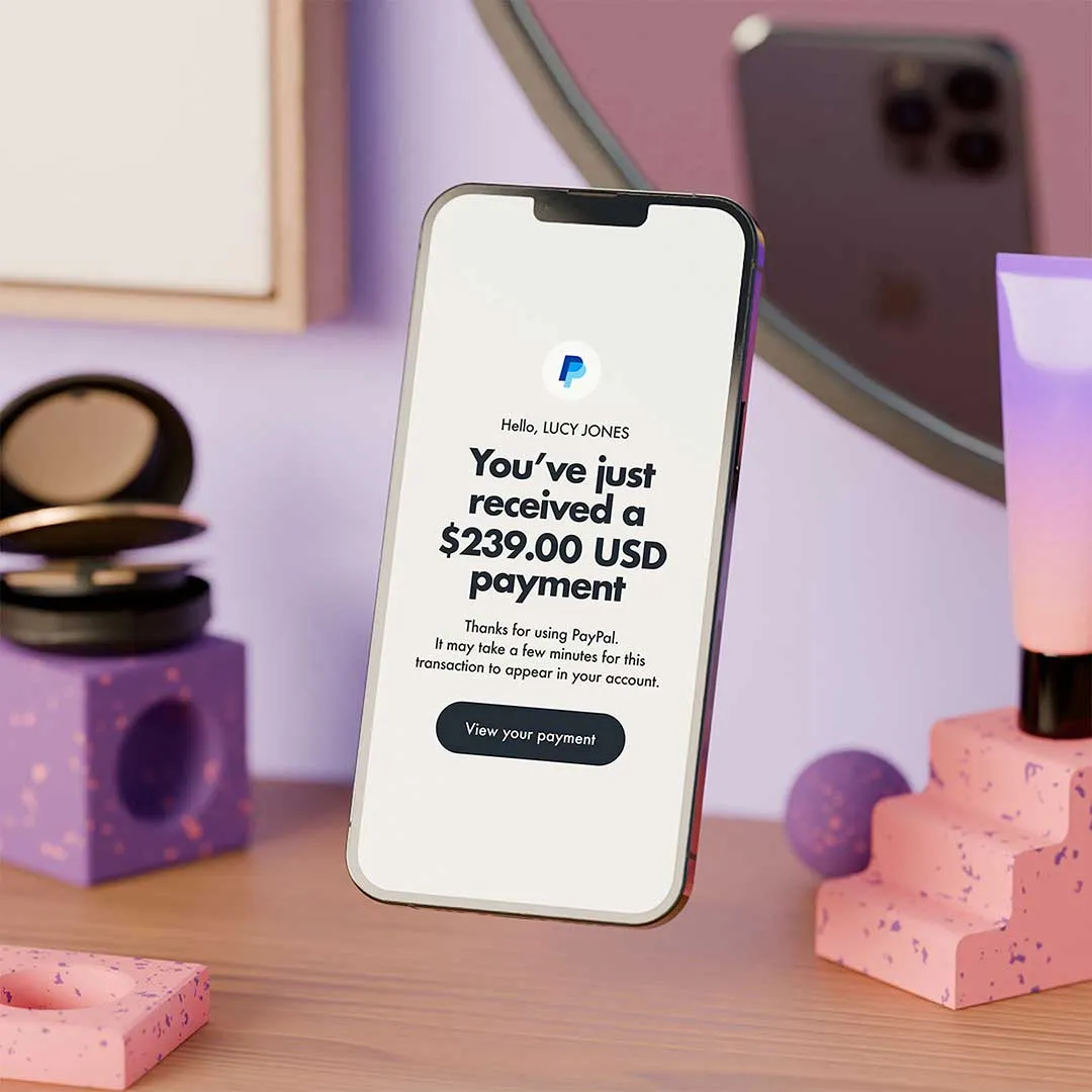 Smartphone displaying a PayPal notification showing a payment, floating above a wooden desk with makeup items and a mirror in the background.
