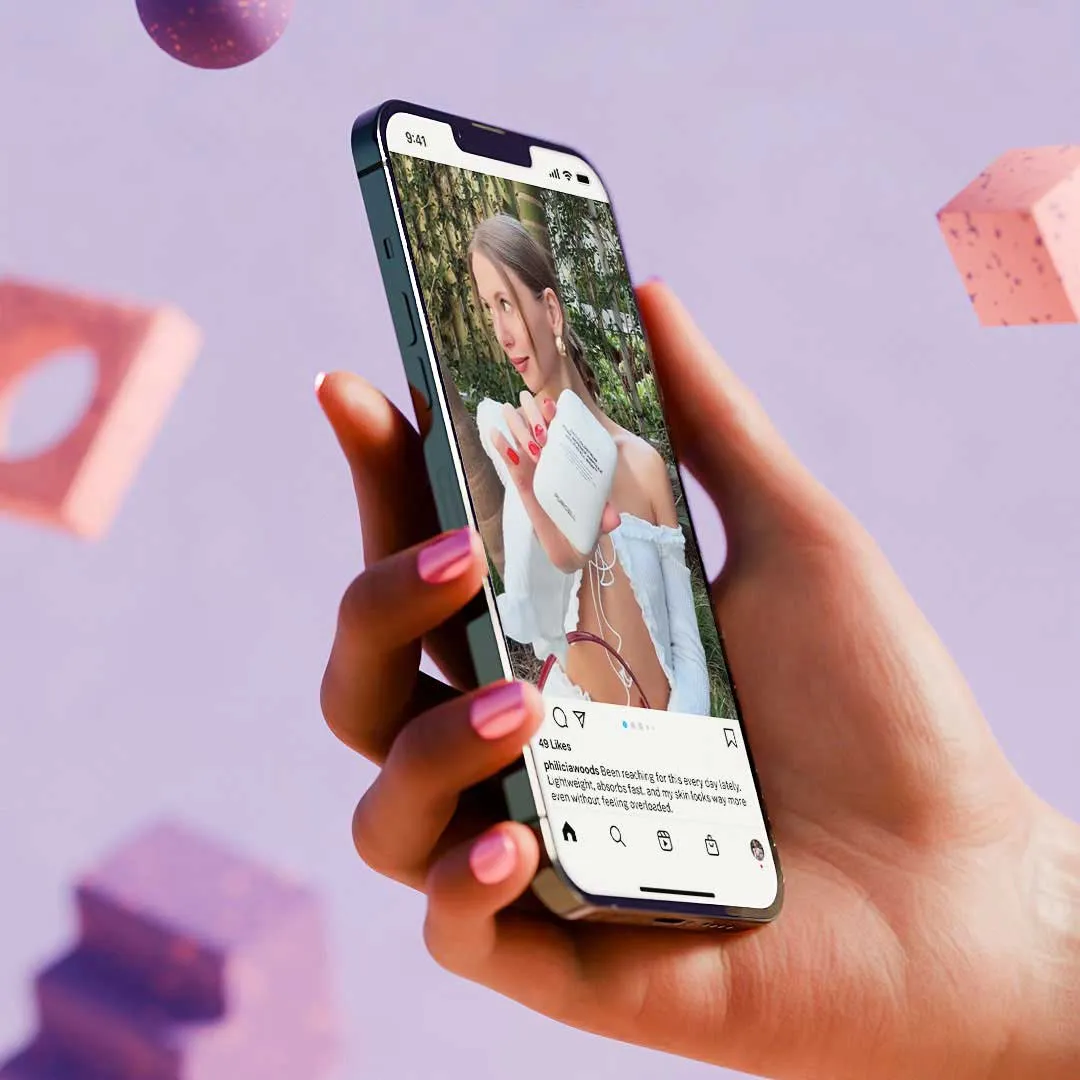 Hand with pink-painted nails holding a smartphone displaying a social media post of a woman in a white off-shoulder top holding a small white device outdoors.