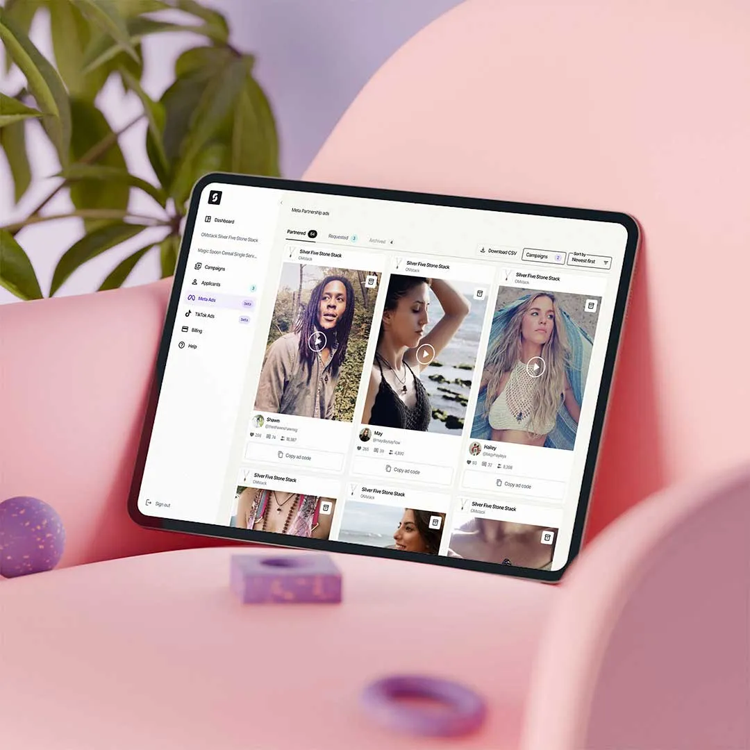 Tablet displaying a Meta Ads dashboard with video thumbnails of diverse individuals, placed on a pink surface with plants in the background.
