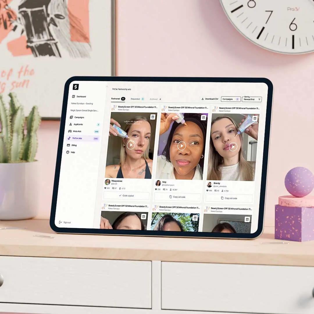 Tablet on desk displaying TikTok Ads platform with video thumbnails of people applying foundation makeup.