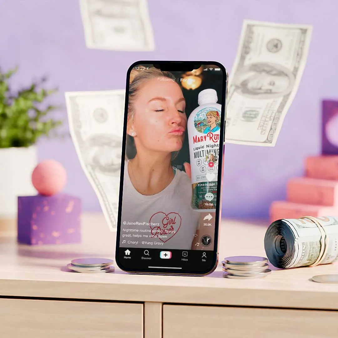 Smartphone on desk shows a TikTok video of a woman holding a product, with flying dollar bills and stacks of coins nearby.