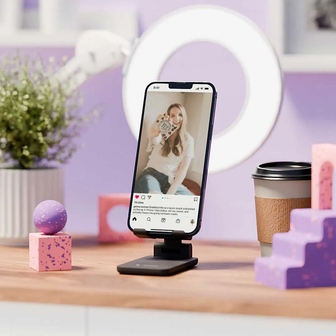 Smartphone on stand showing a smiling woman holding a product, with a ring light, coffee cup, and decorative objects in the background.