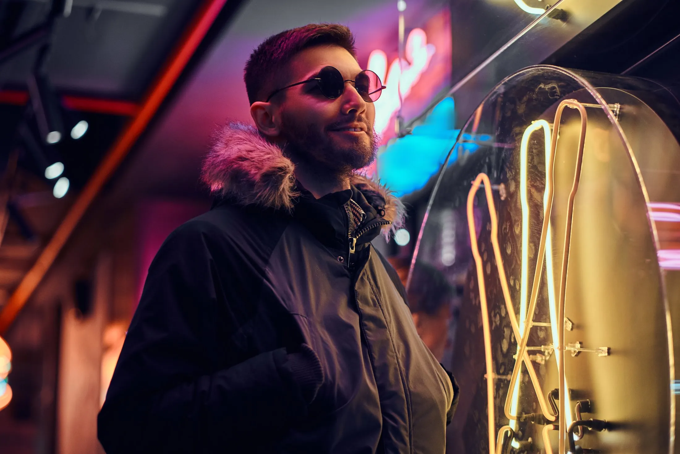 Person in sunglasses and fur-trimmed jacket standing near neon lights