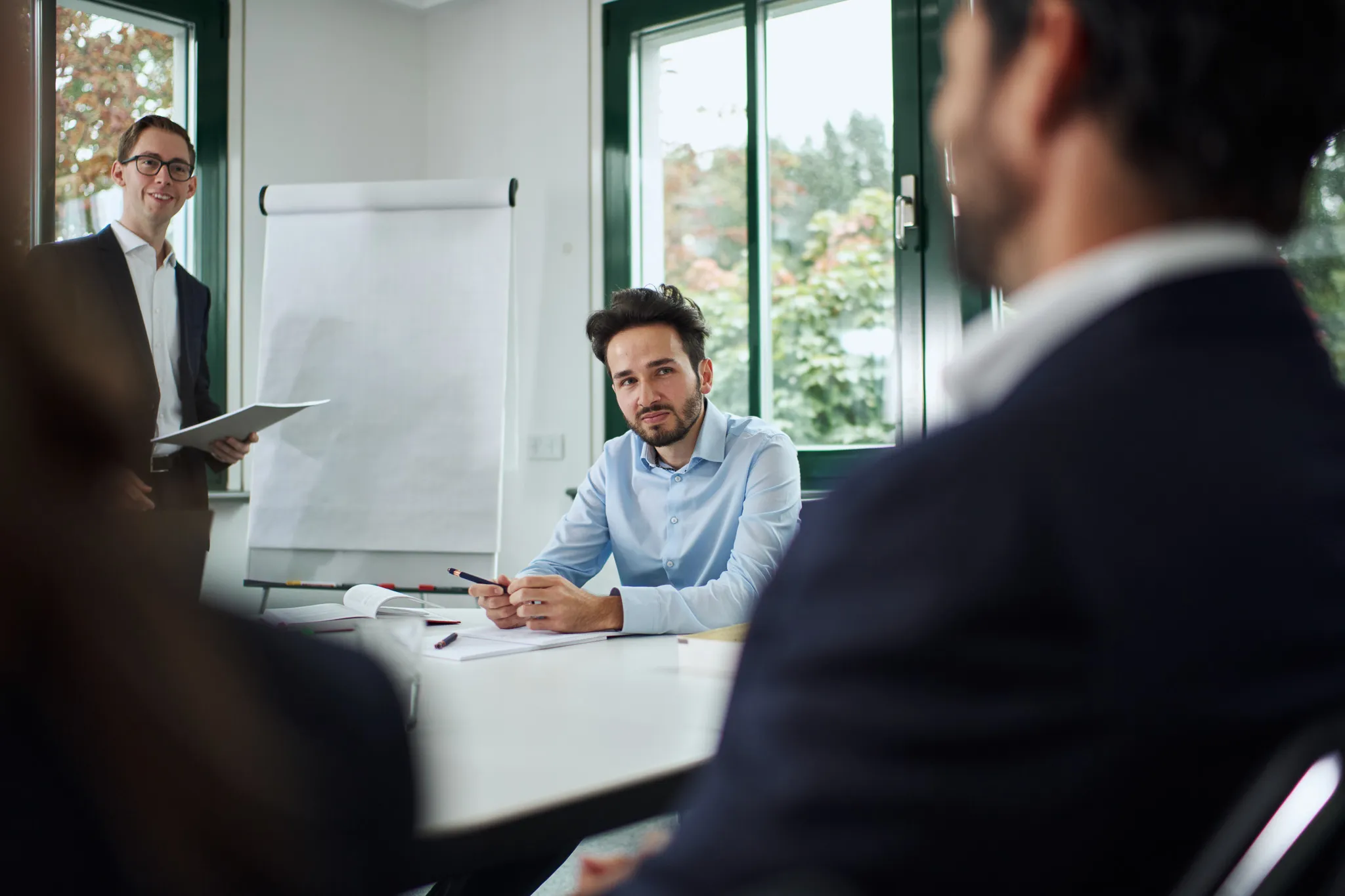 Mann in blauem Hemd hört aufmerksam bei einem Meeting mit einem Kollegen, der vor einem Flipchart steht.
