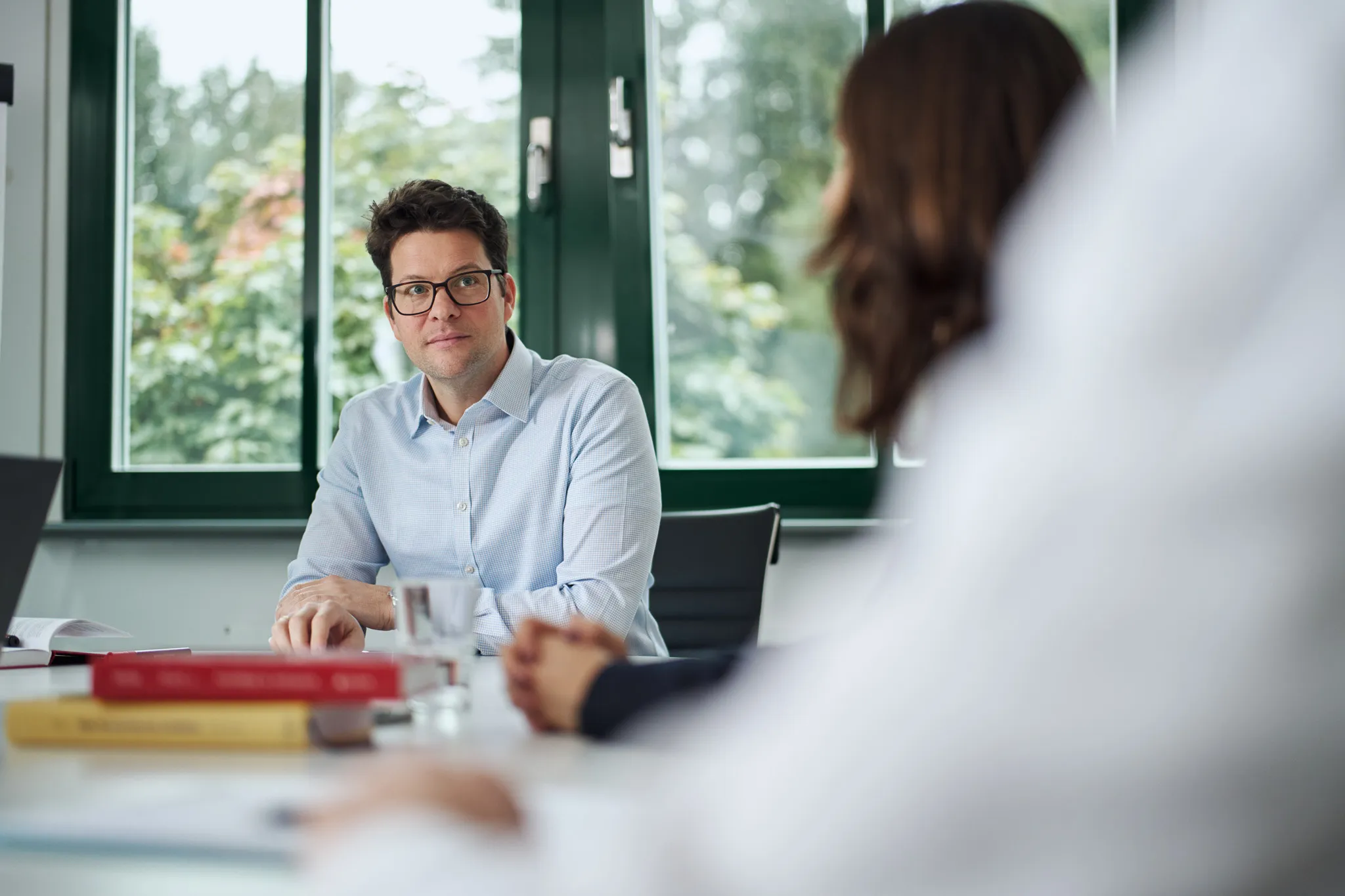 Ein Mann mit Brille und blauem Hemd sitzt an einem Tisch im Büro und hört aufmerksam zu.