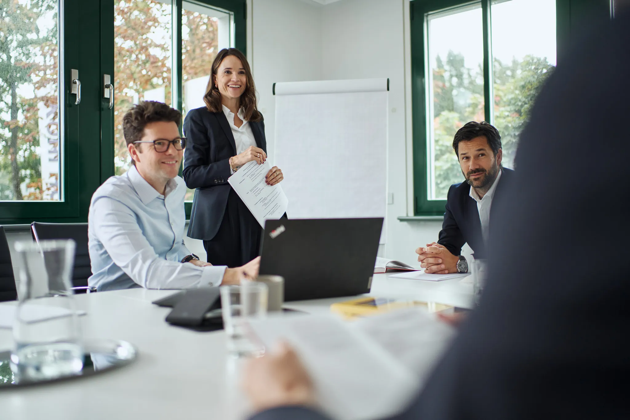 Drei Geschäftsleute in einem modernen Büro, eine Frau steht mit Dokumenten vor einem Flipchart, zwei Männer sitzen am Tisch mit Laptop.