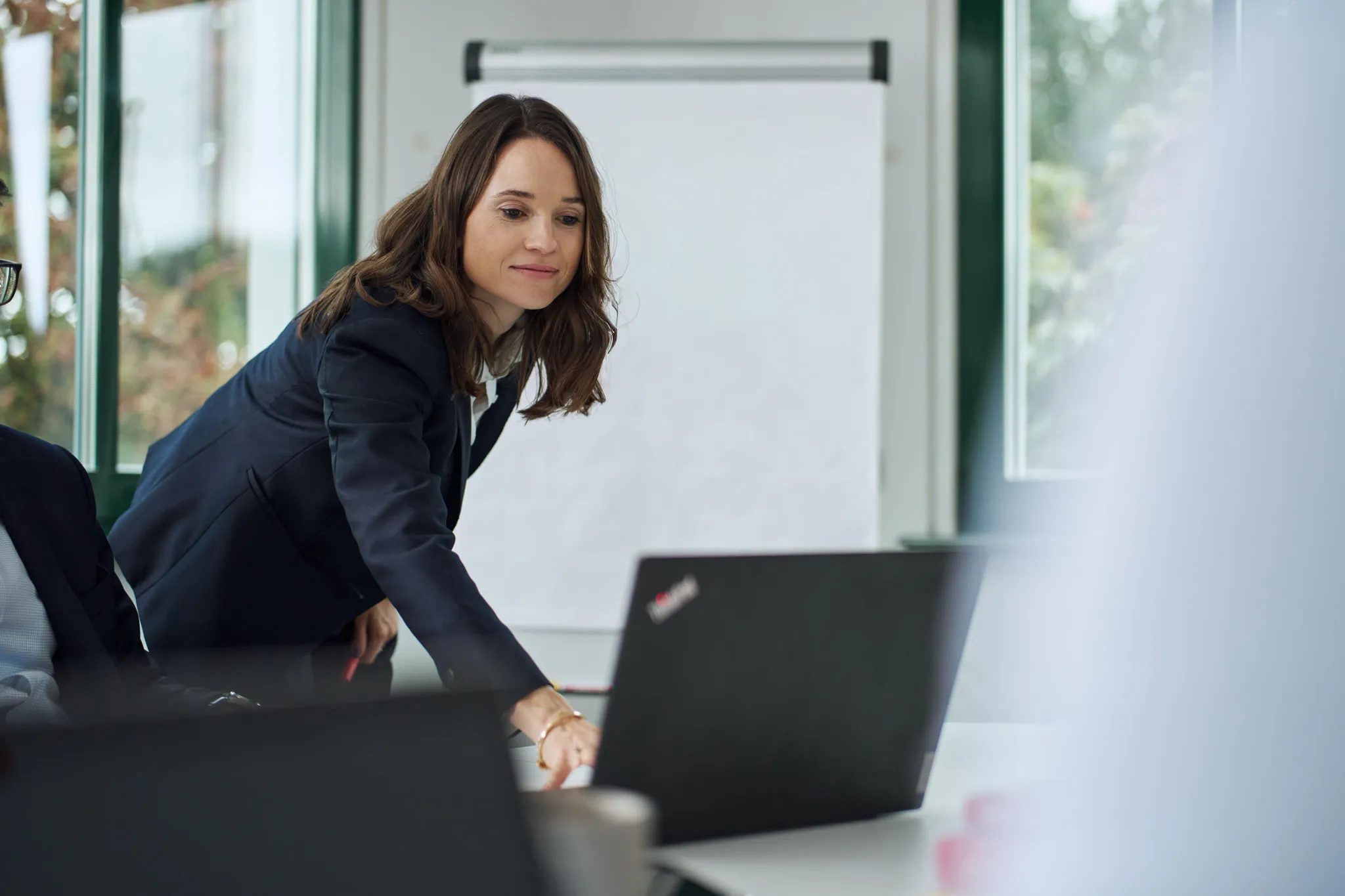 Geschäftsfrau zeigt mit dem Finger auf einen Laptop in einem modernen Büro mit Flipchart und großen Fenstern.