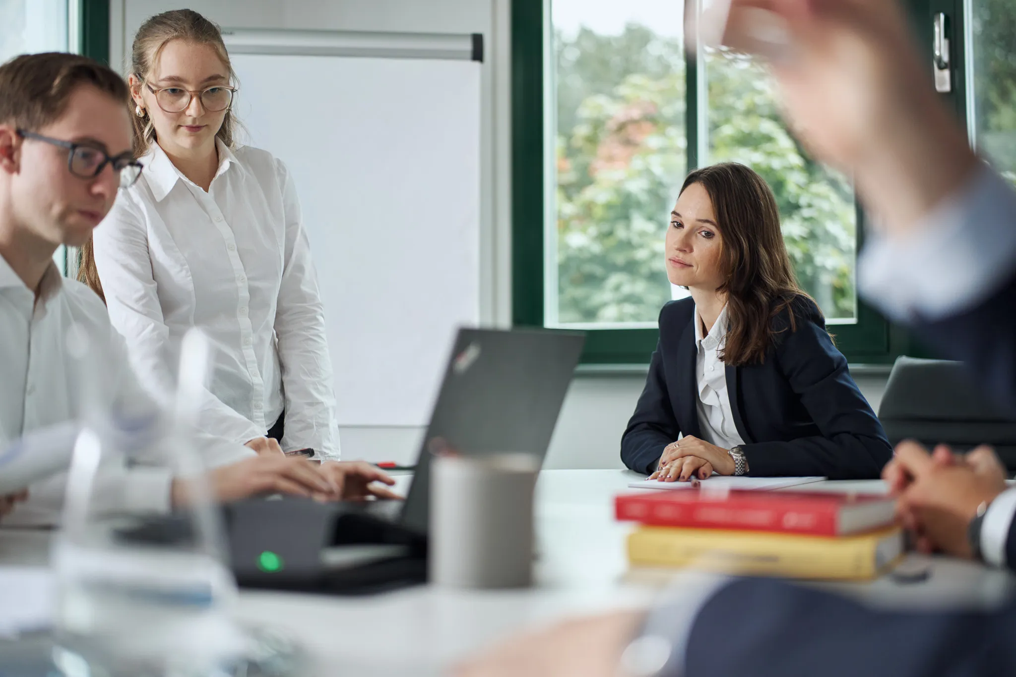 Menschen in formeller Kleidung sitzen in einem modernen Besprechungsraum mit einem Laptop und Büchern auf dem Tisch.