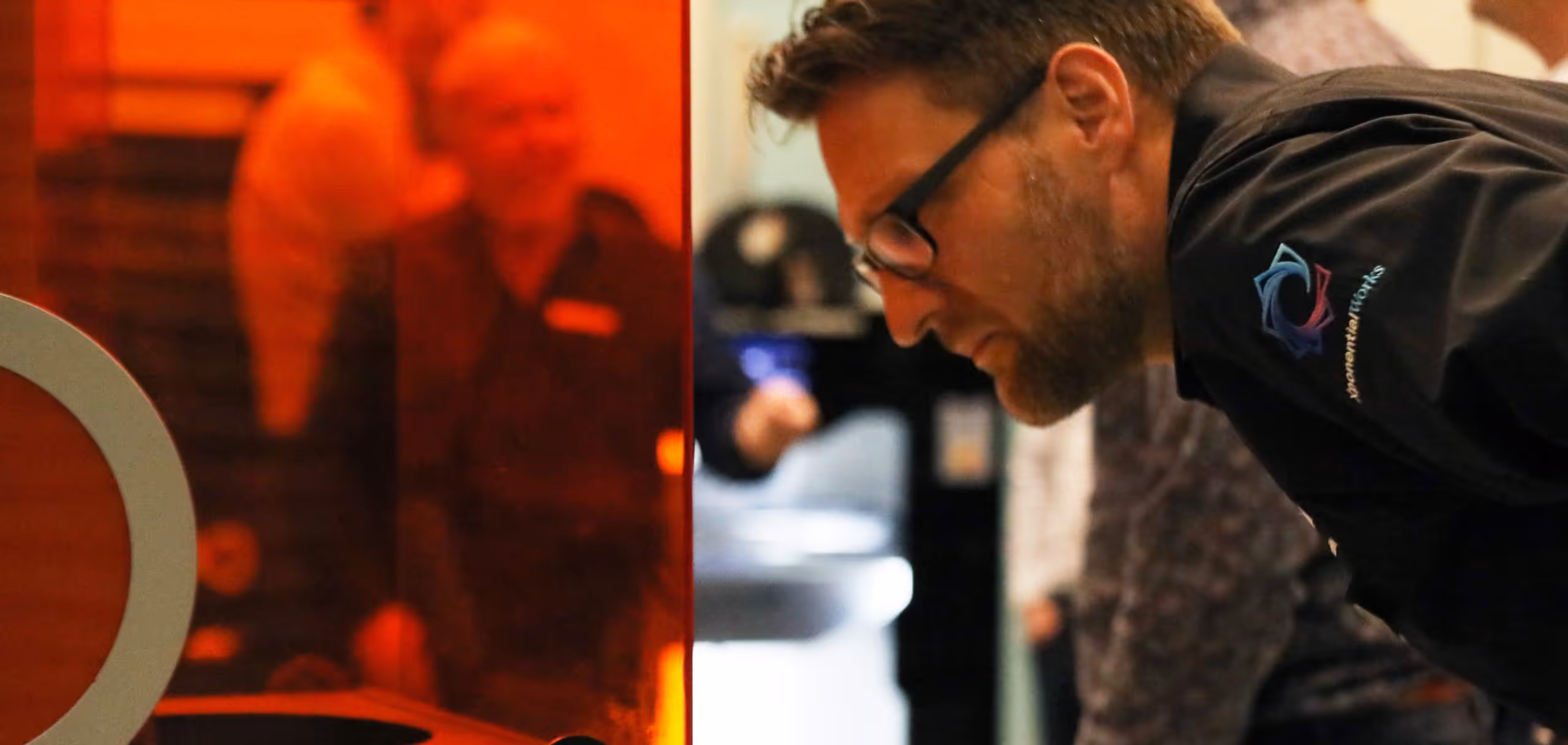 A young woman working in a machine shop. She is looking directly towards the camera, and is surrounded by men.