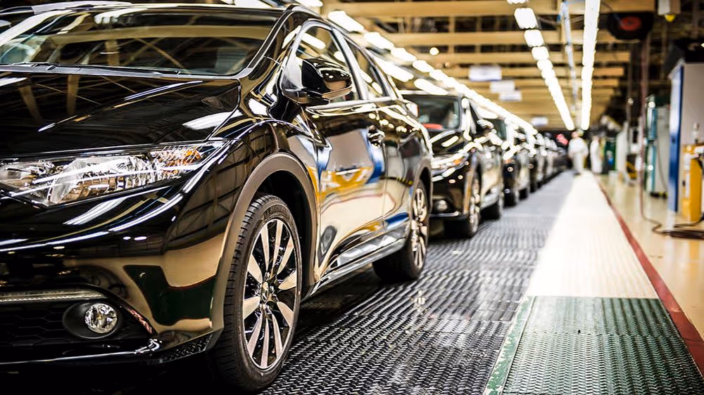 A young woman working in a machine shop. She is looking directly towards the camera, and is surrounded by men.