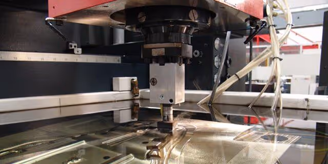 A young woman working in a machine shop. She is looking directly towards the camera, and is surrounded by men.