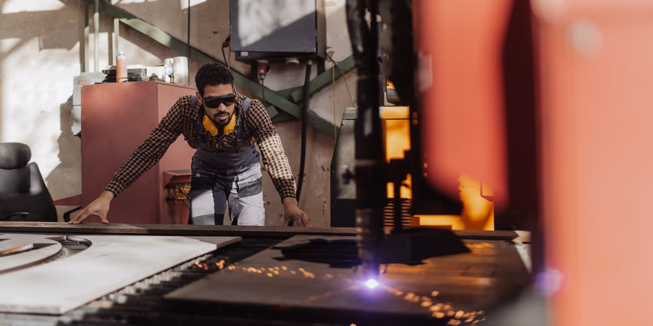 A young woman working in a machine shop. She is looking directly towards the camera, and is surrounded by men.