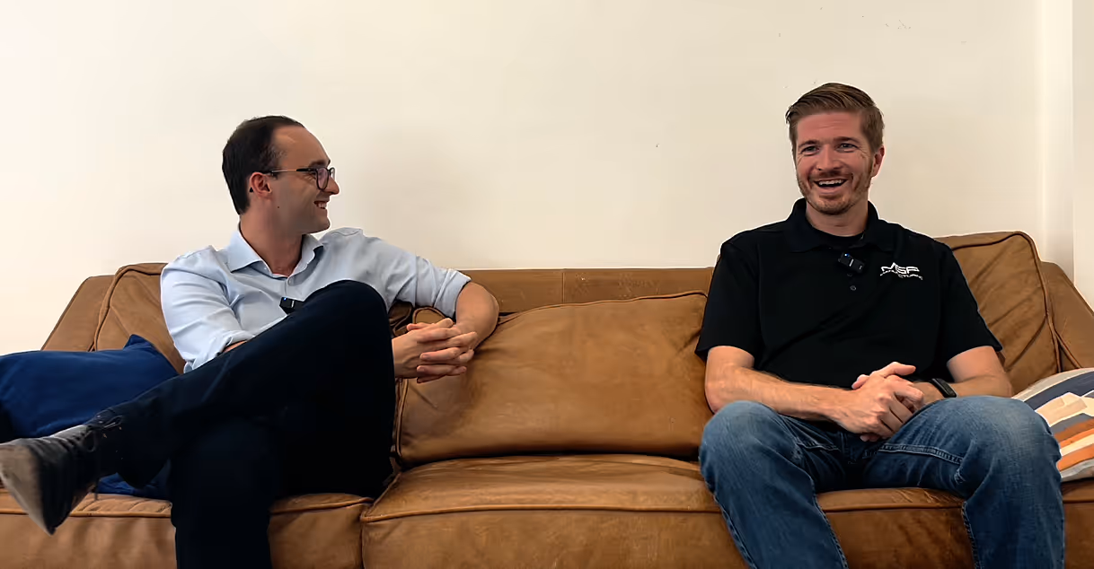Two men sitting on a sofa. On the left is Henry Morris, on the right is Johnny Goode. They are smiling, and mid conversation.