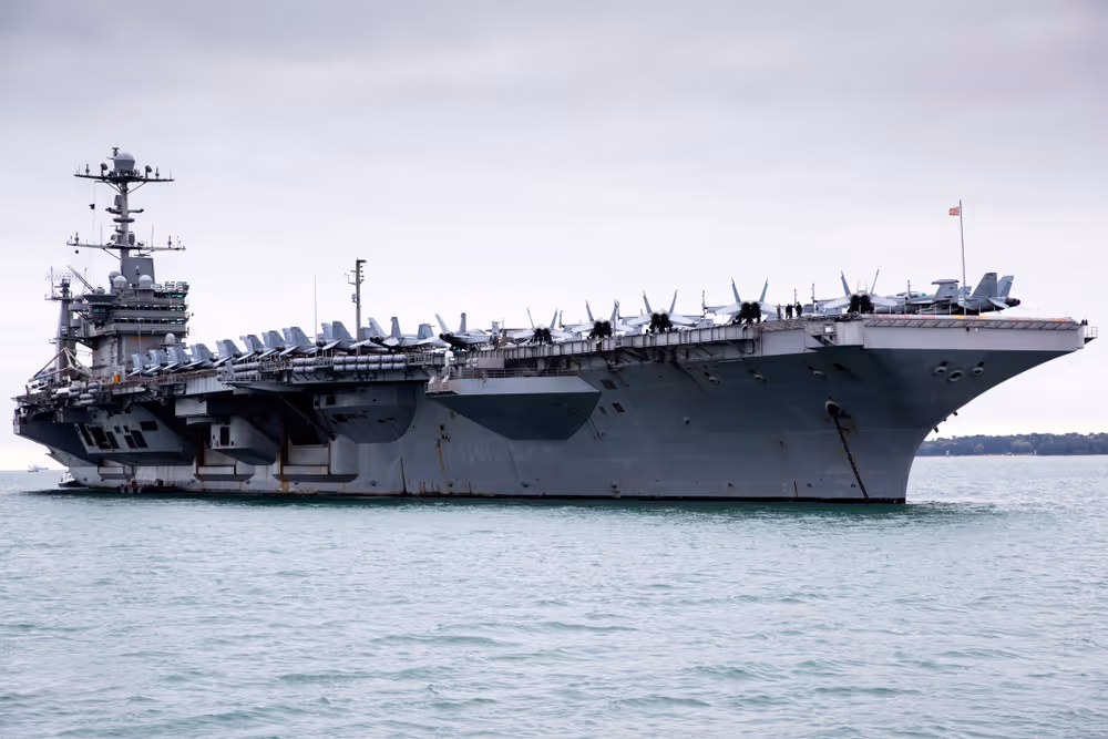 A young woman working in a machine shop. She is looking directly towards the camera, and is surrounded by men.
