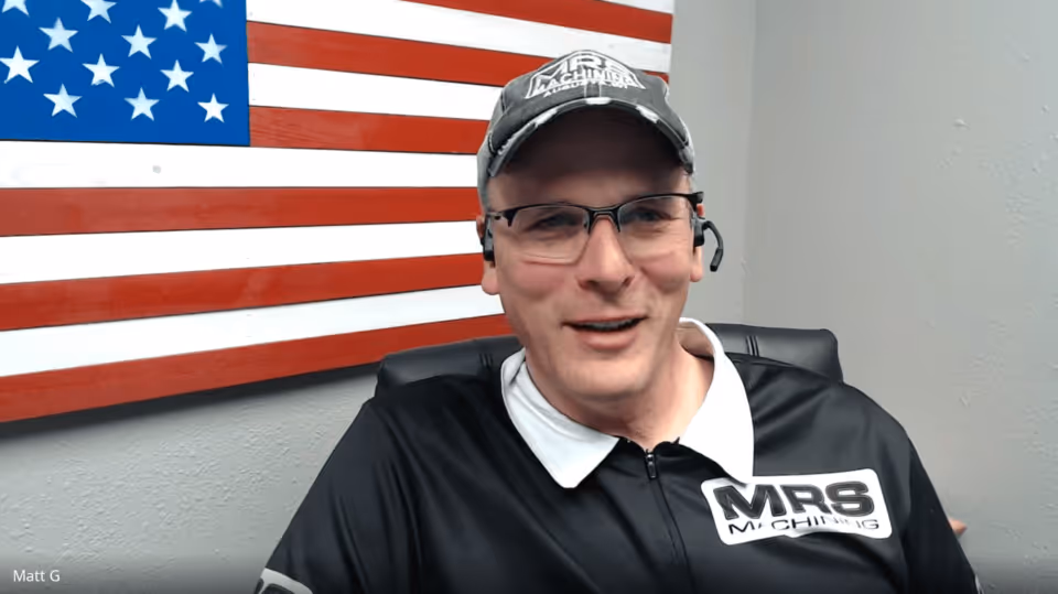 A picture of Matt Guse. He is looking into the camera and smiling. Behind him is the American flag. He is wearing a black t-shirt with a white collar, and a black baseball hat, both of which have MRS Machining stamped onto them.