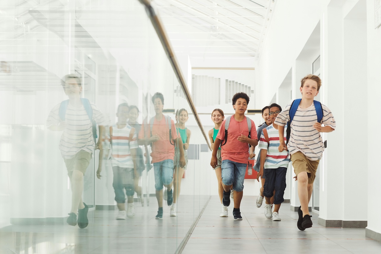Group of diverse children running down a bright school hallway with backpacks.