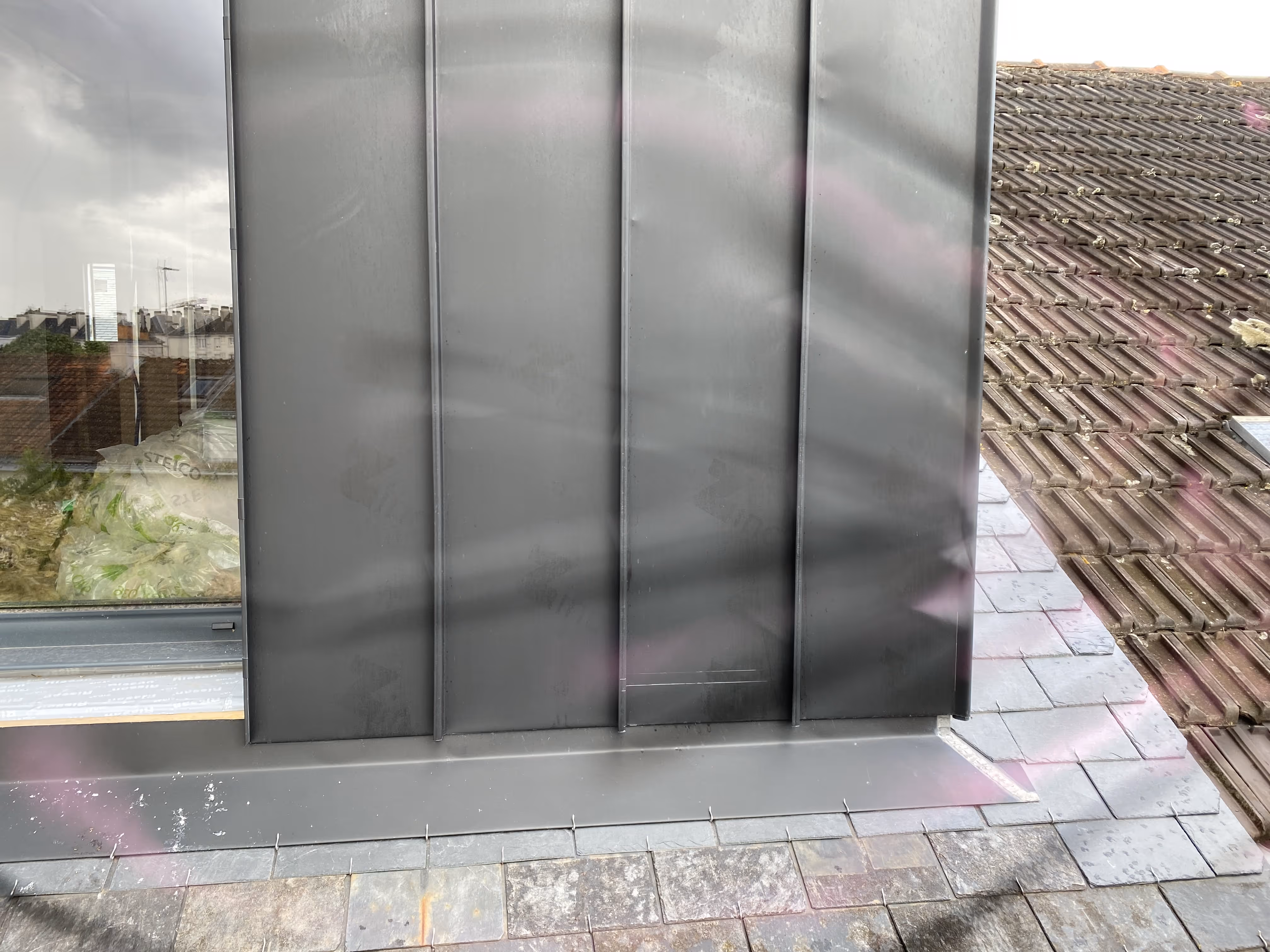 Vue rapprochée d'une partie de toit avec une fenêtre, des tuiles anciennes à droite, et une surface métallique plate grise au centre.