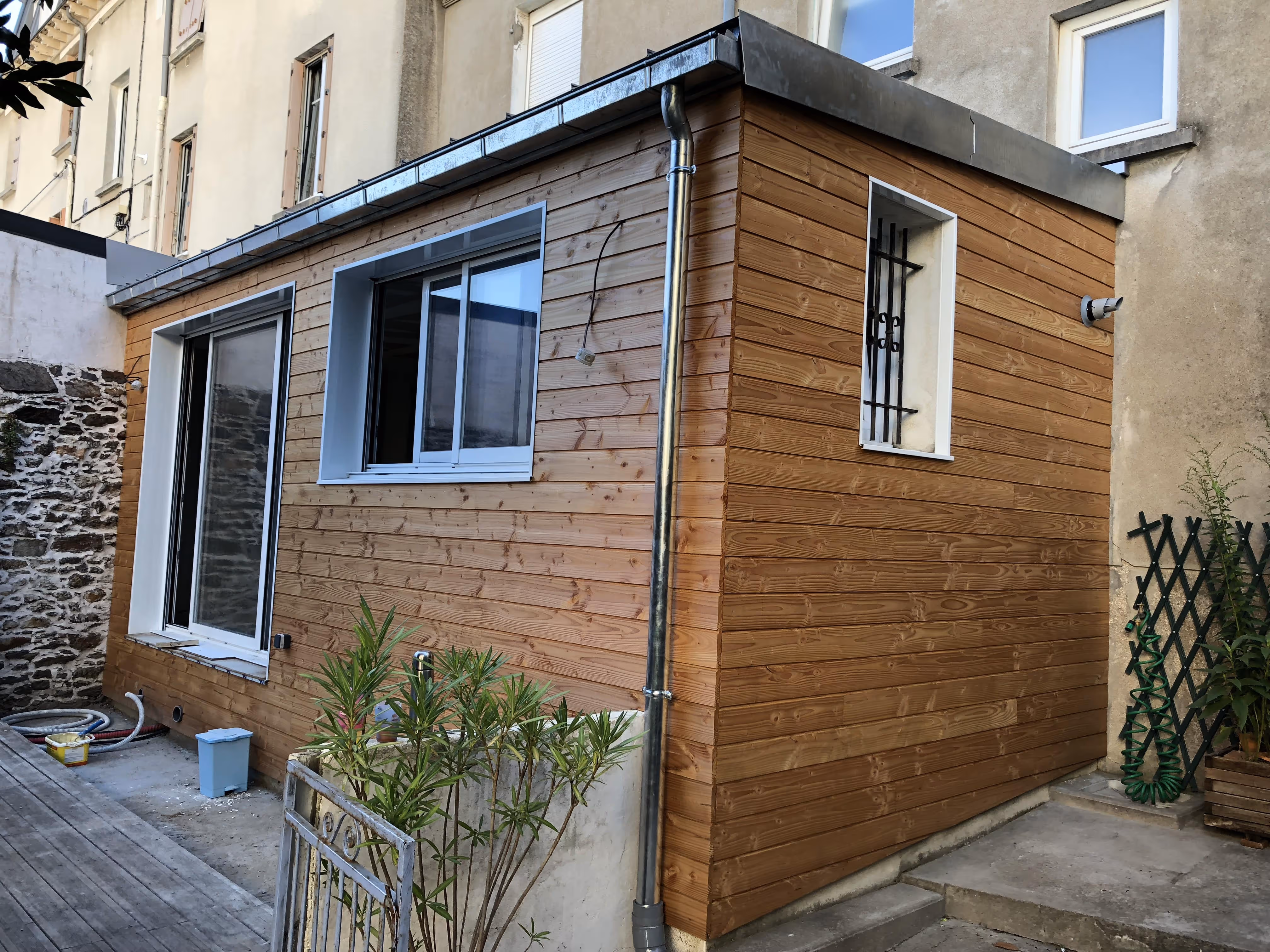Petite extension de maison en bois avec deux fenêtres, une grille de protection et un tuyau de descente d'eau.
