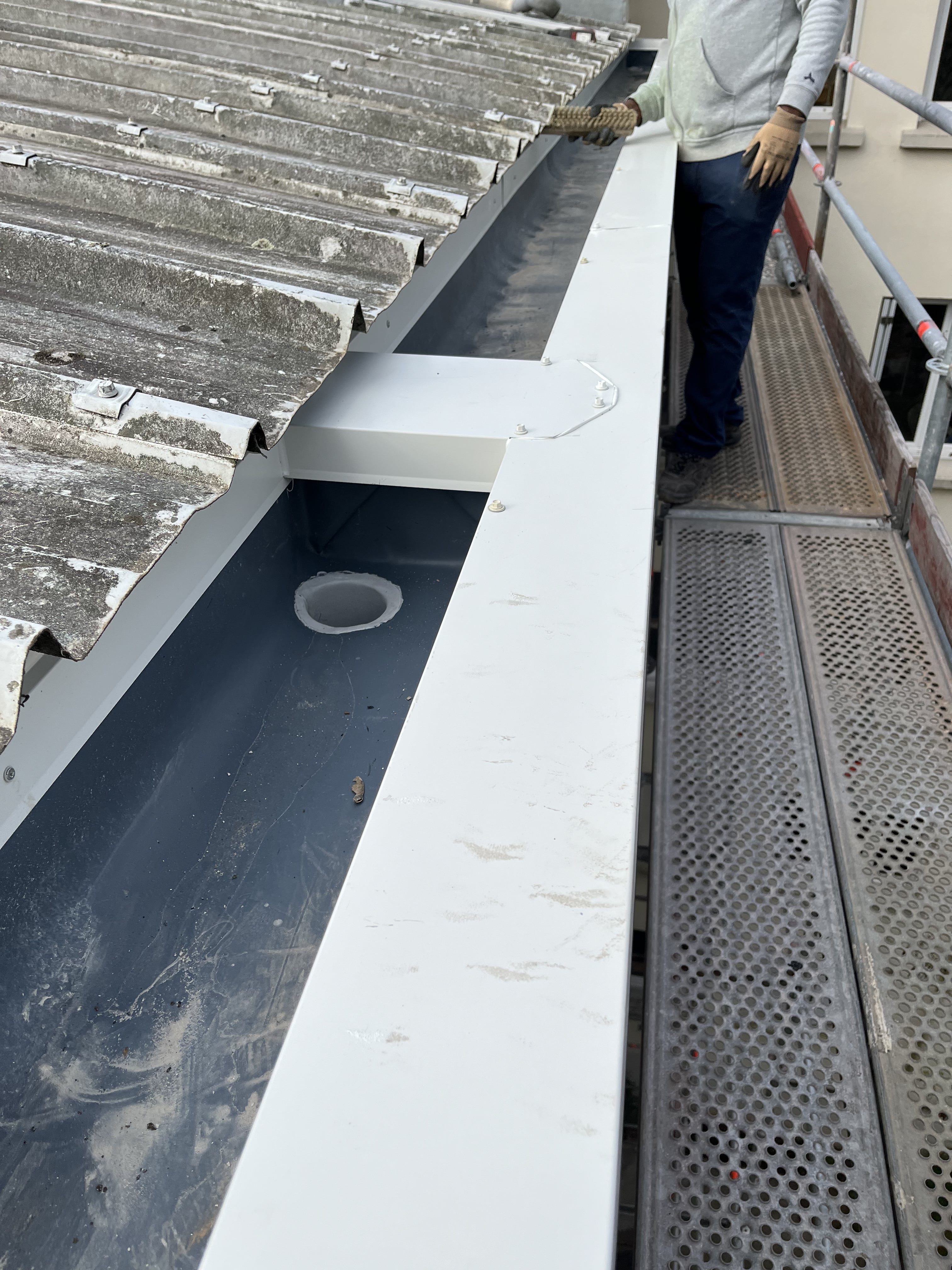 Travailleur portant des gants inspectant une gouttière propre en métal sous un toit en plaques ondulées grises sur un échafaudage.