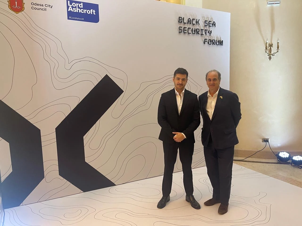 Two people in suits without ties pose for a photo in front of the stand