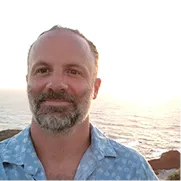 Man with gray beard and patterned shirt photographed at sunset by the ocean