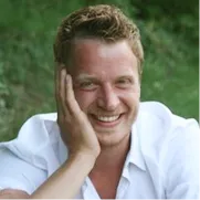 Smiling man with wavy blonde hair in a white shirt, photographed outdoors