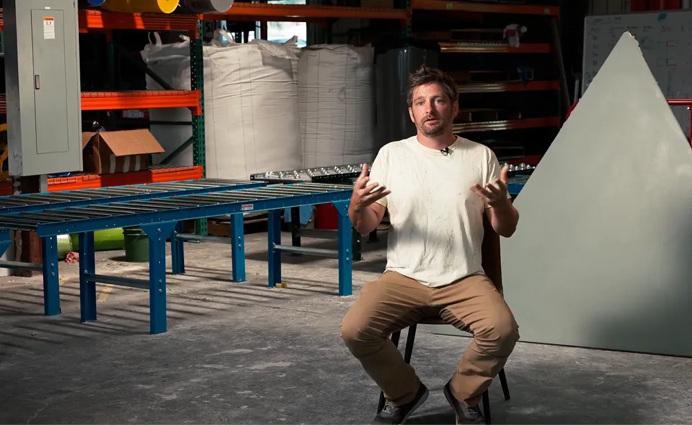 Geoship founder speaking in the factory with ceramic dome panel and conveyor belt visible in the background