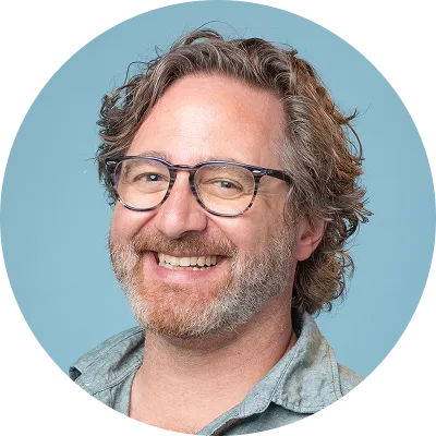Smiling man with curly hair and glasses wearing a gray button-up shirt