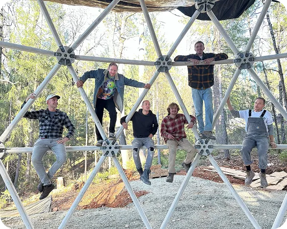 Six Geoship team members sitting casually on the geometric frame of a dome under construction