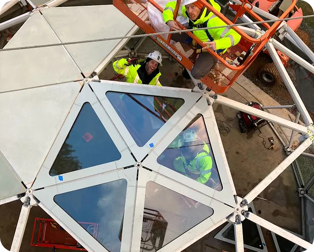 Geoship workers in hard hats and safety vests installing triangular ceramic panels on a dome using a lift