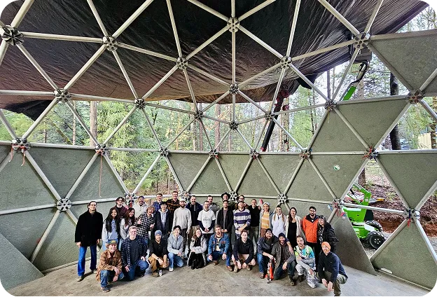 Geoship team group photo inside a partially constructed geodesic dome frame in a forested setting