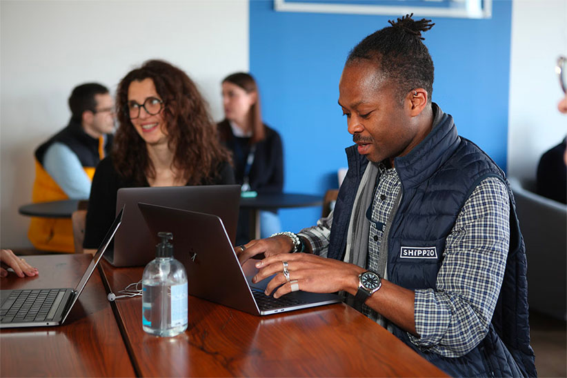 Group of five young professionals collaborating with laptops, tablets, and notebooks in a modern office space.