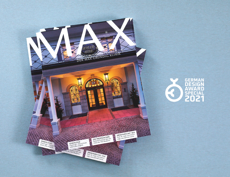 Stack of MAX health magazine issues showing an illuminated entrance of a building on the cover, next to the German Design Award Special 2021 logo on a blue background.