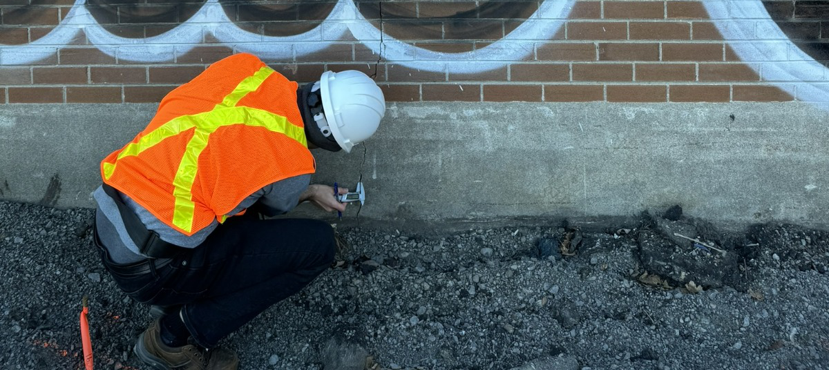 Engineer performing a structural inspection on-site, kneeling near a brick wall to examine the building foundation.