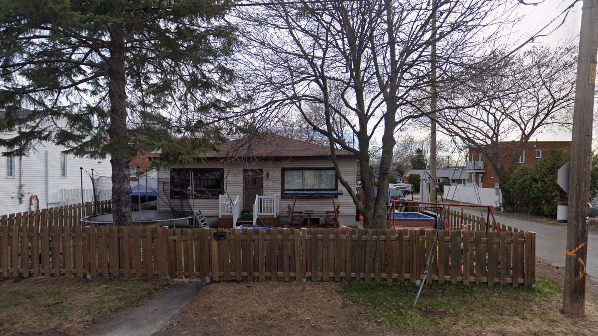 Maison à un étage avec clôture en bois, trampoline et balançoire dans la cour arrière sous un ciel nuageux.