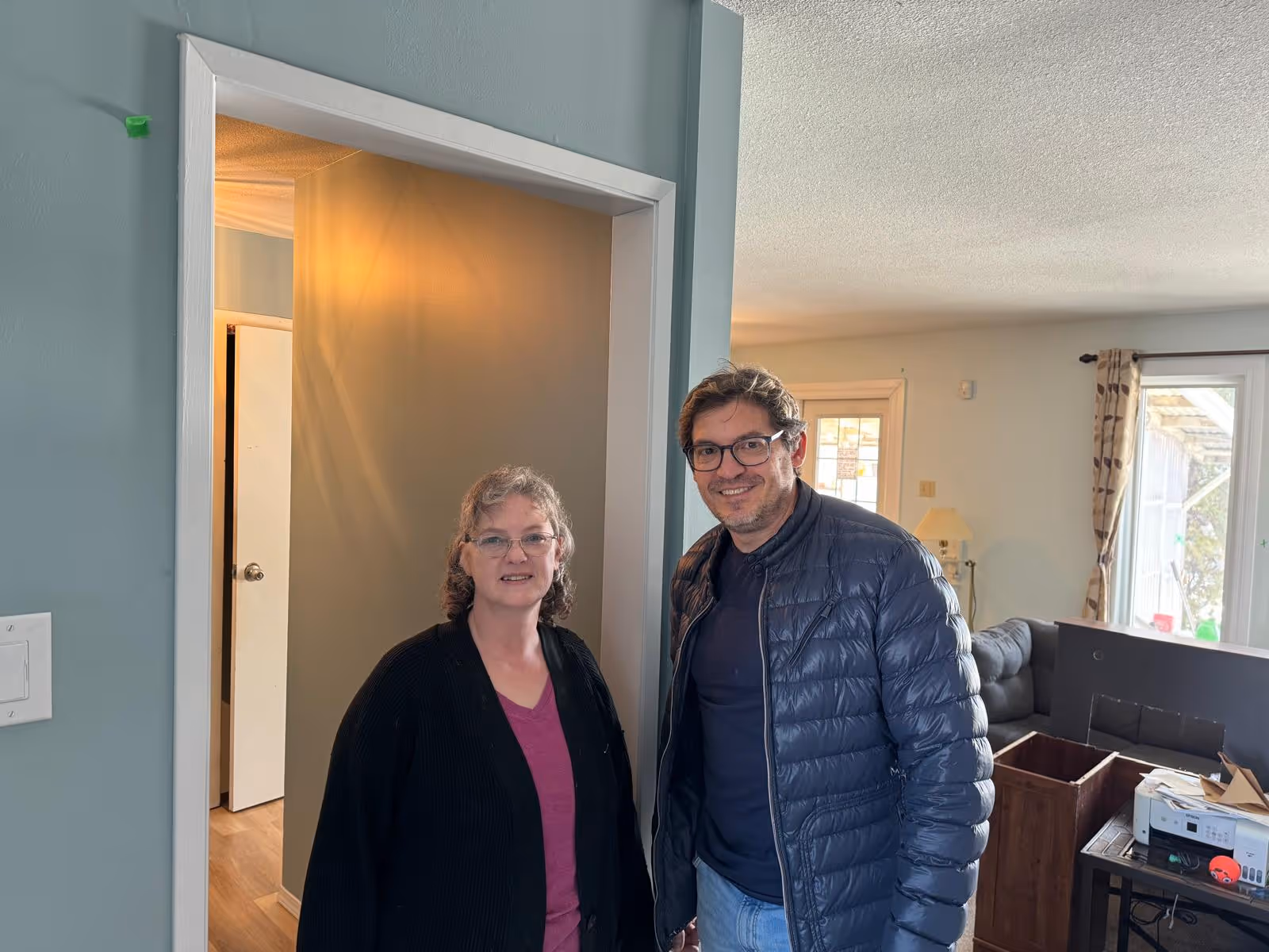Un homme et une femme souriants posent à l'intérieur d'une maison près d'une porte entrouverte.