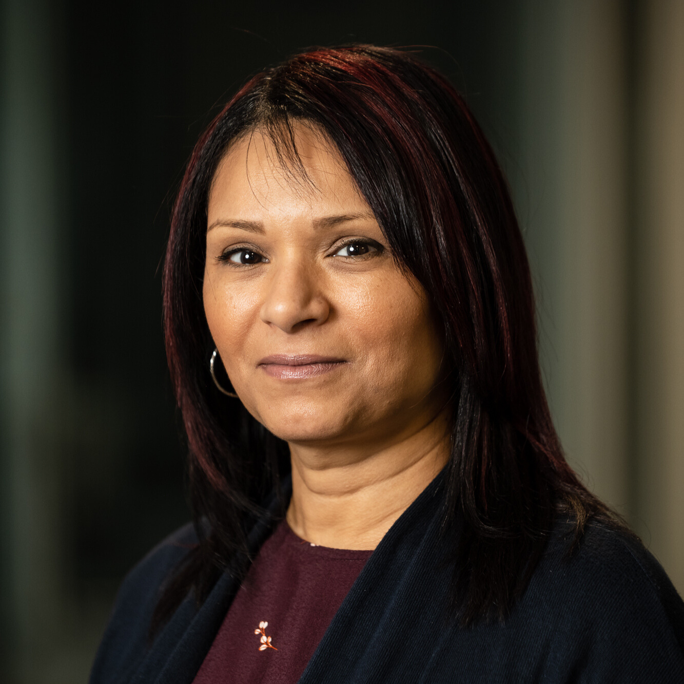 Portrait of a woman with straight dark hair wearing a maroon top and black cardigan, smiling slightly against a blurred dark background.