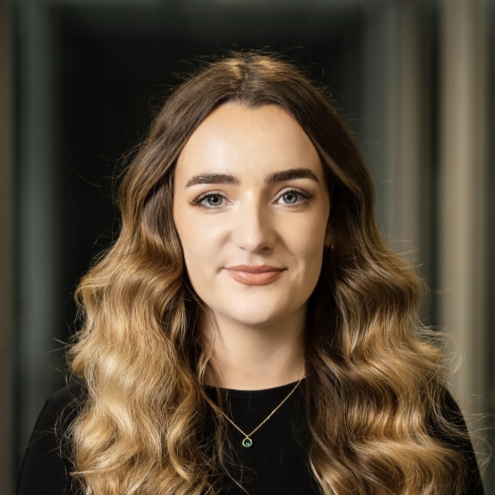 Young woman with long wavy light brown hair, wearing a black top and a gold necklace with a green pendant, smiling softly against a blurred dark background.