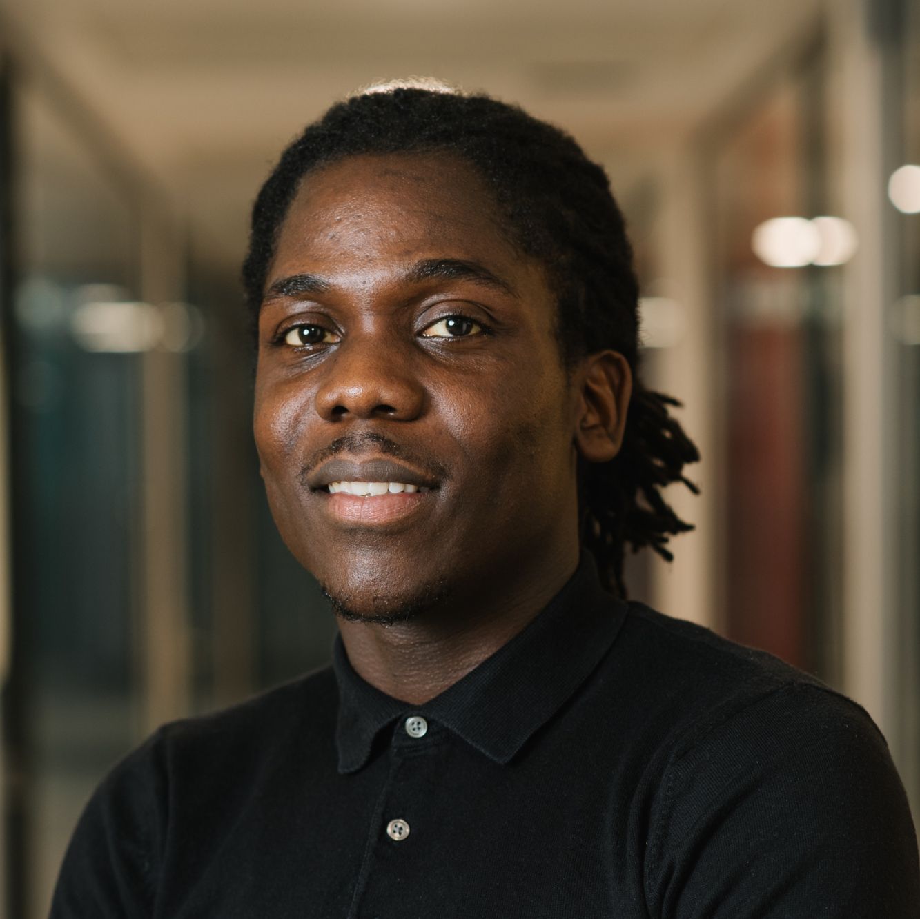 Smiling young Black man with dreadlocks wearing a black collared shirt in an indoor setting.