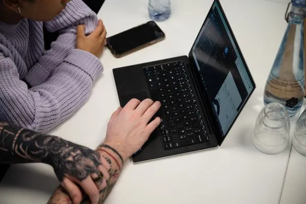 Two people looking at a laptop screen on a white table, with a smartphone, water bottle, empty glasses, and one person's tattooed arm visible.