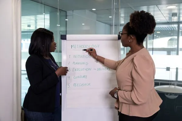 Two professional women discuss a flip chart listing project methodology steps: initiation, planning, execution, closure, and feedback in a modern office.