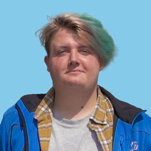 Charlie has short, styled hair wearing a blue jacket over a yellow checkered shirt against a blue background.