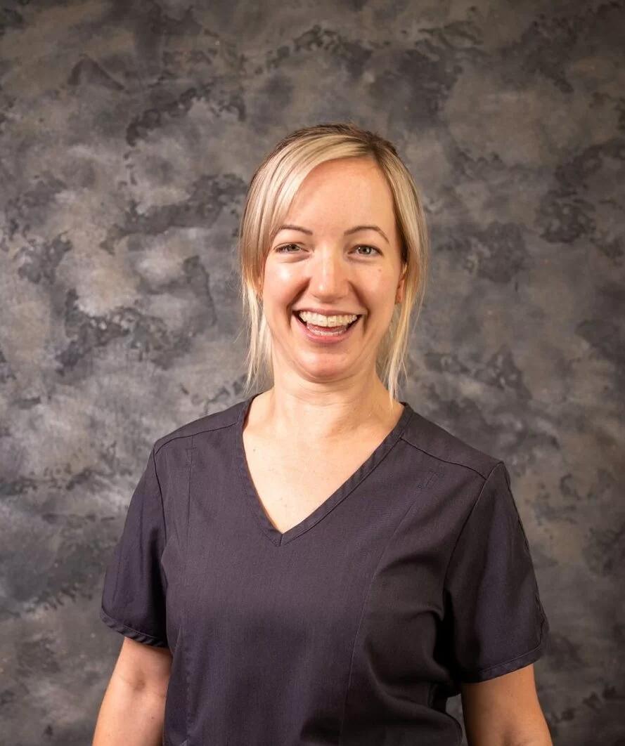 Smiling blonde woman in a dark gray scrub top standing against a mottled gray background.