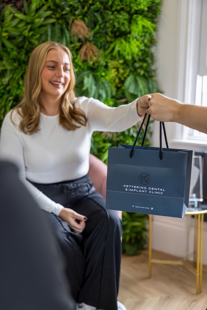 Smiling woman in white top sitting while receiving a navy blue shopping bag labeled 'Kettering Dental & Implant Clinic' from another person.