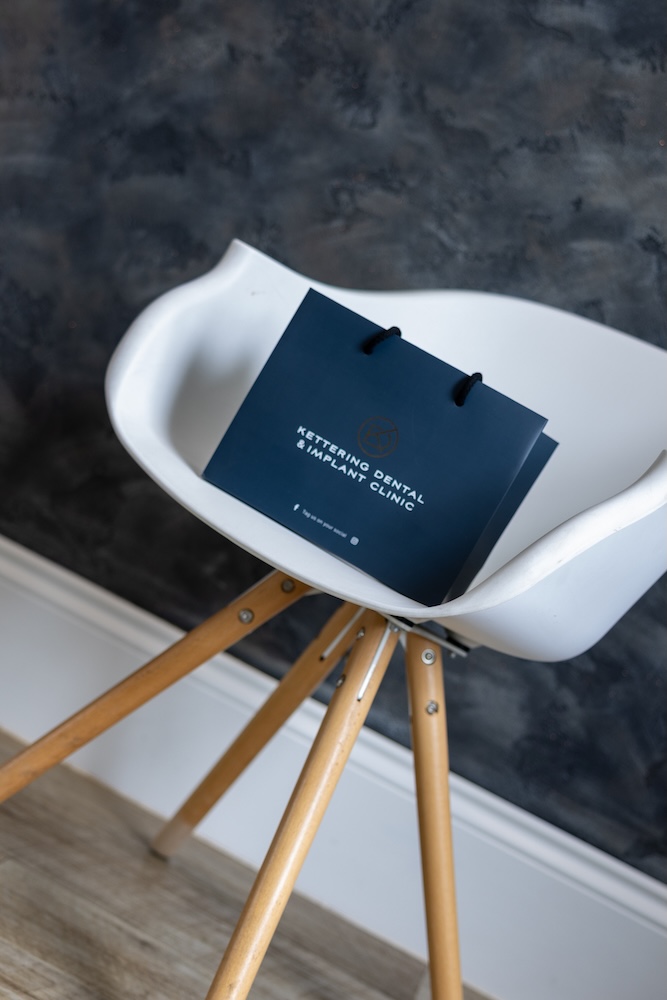 Navy blue Kettering Dental & Implant Clinic bag resting on a white chair with wooden legs against a textured dark wall.