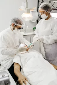 2 Dentists wearing their professional white uniforms and hair cap, cleaning a patient's teeth.