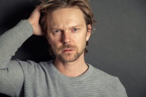Confused man wearing a grey sweatshirt, holding his hear with his right hand, thinking of going for a dental check up
