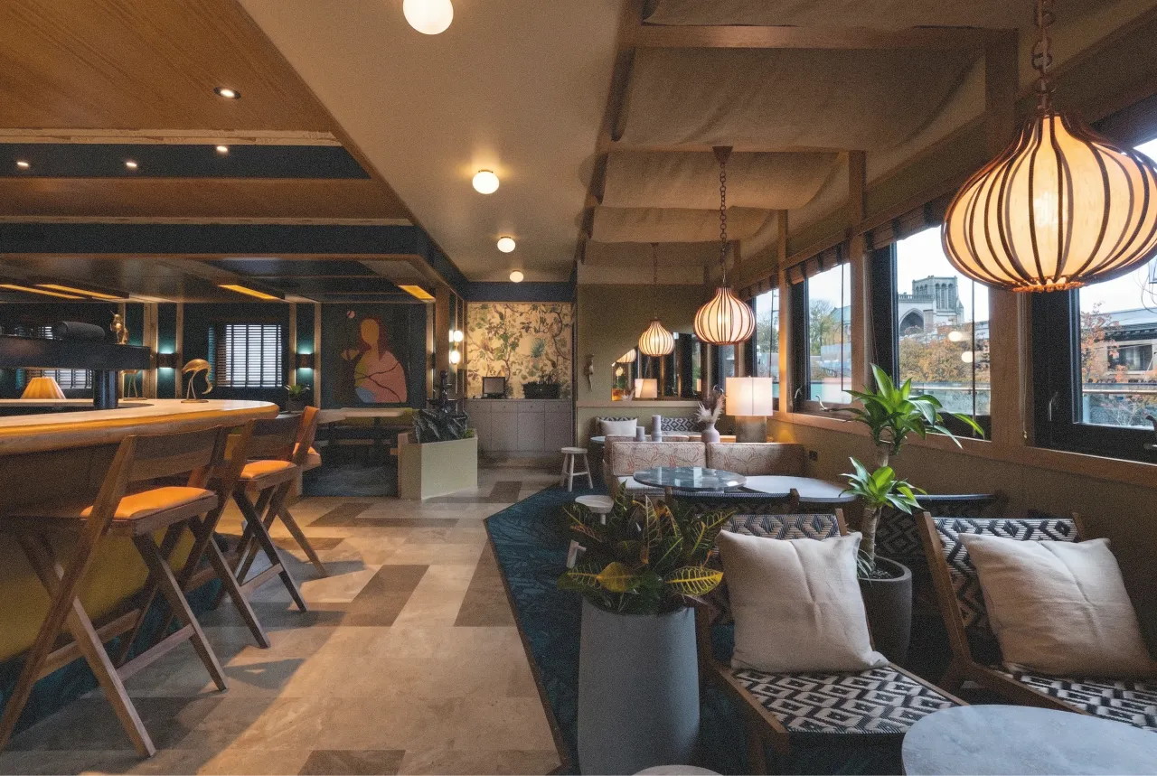 Cozy lounge area with patterned chairs, cushions, pendant lights, and large windows with city view. Superflux Cabana Edda Construction.