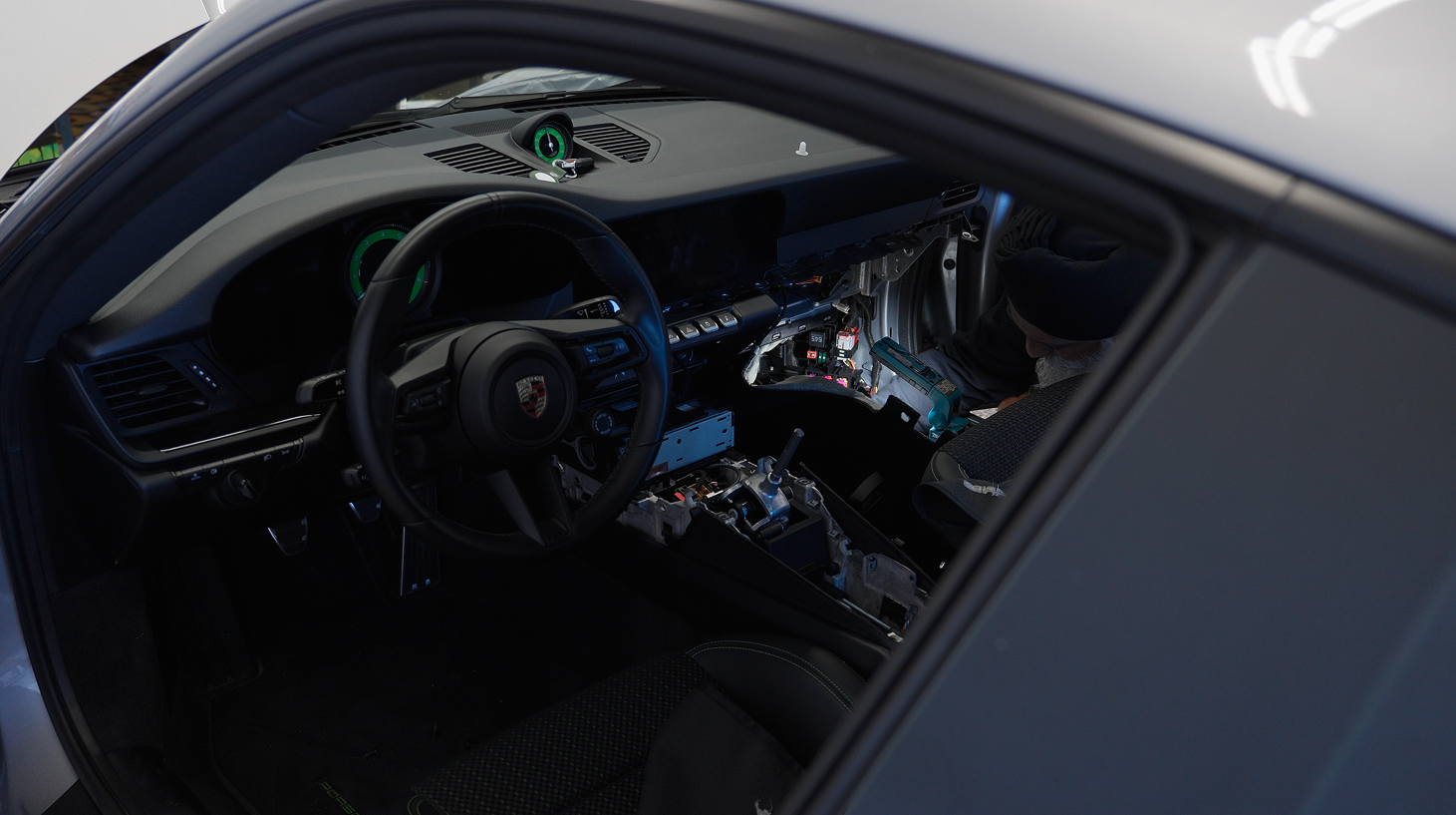 Man working on dismantled car dashboard with steering wheel showing Porsche emblem.