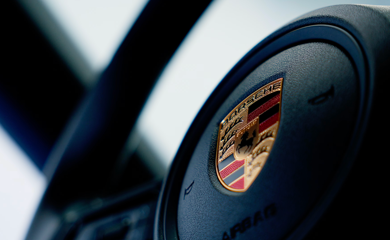 Close-up of a Porsche steering wheel with logo in the center on a black textured background.